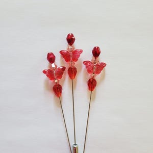 May include: Three decorative hat pins with red butterfly and bead designs. Each pin features a red butterfly, red beads, and a silver-colored pin. The pins are arranged against a white background.