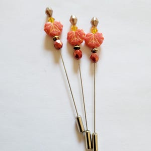 May include: Three decorative hat pins with leaf-shaped beads in shades of orange and red. Each pin features a silver-colored shaft, a faceted bead, and a metallic bead at the top. The pins are arranged on a white background.