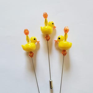 May include: Three yellow duck pins with orange accents. Each pin features a small, glossy yellow duck with an orange beak and a decorative orange bead. The pins have silver-colored shafts and cylindrical tips. The pins are set against a white background.