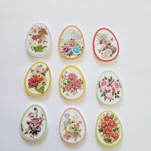 May include: Nine decorative Easter egg-shaped ornaments with floral designs. Each egg features a different arrangement of flowers in various colors, including pink, red, and yellow, set against a white background. The edges are trimmed with different colored borders.