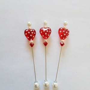 May include: Three decorative hat pins. Each pin features a red heart-shaped bead with white polka dots, a small red bead, a pearl bead, and a silver-colored pin. The pins have a large white pearl at the base.