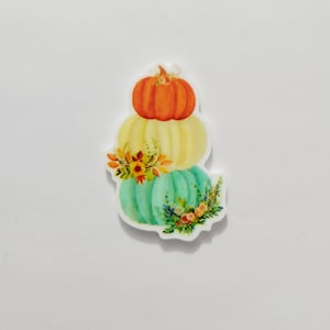 May include: A stack of three pumpkins with fall-themed floral accents. The top pumpkin is orange, the middle pumpkin is yellow, and the bottom pumpkin is teal.
