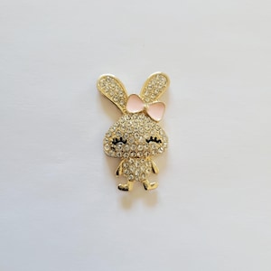 May include: A gold-toned bunny brooch encrusted with clear rhinestones. The bunny has a pink bow and black eyes. The brooch is a cute and sparkly accessory.