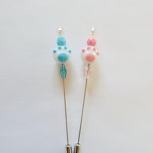 May include: Two decorative hat pins. One pin features a blue paw print design with blue and clear beads. The other pin has a pink paw print design with pink and clear beads. Both pins have silver-colored metal pins.