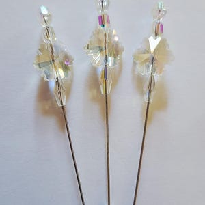 May include: Three decorative hat pins with iridescent crystal accents. Each pin features a silver-toned metal shaft and a faceted crystal design. The crystals reflect light, creating a sparkling effect. The pins are arranged against a white background.