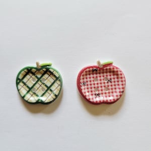 May include: Two apple-shaped buttons. One is green with a plaid pattern, and the other is red with a gingham pattern. Both have a small green stem and are made of ceramic or a similar material.