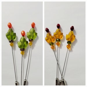 May include: A set of six decorative hat pins. Three pins feature green glass leaf designs with orange bead accents. The other three pins have amber glass leaf designs with burgundy bead accents. All pins have silver-toned metal shafts.