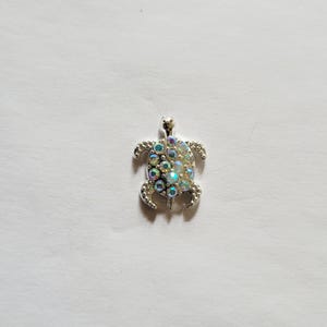 May include: A silver-toned turtle pendant with iridescent, multi-colored rhinestones. The pendant has a loop at the top for a chain and is set against a white background. The turtle's shell is covered in the sparkling stones.