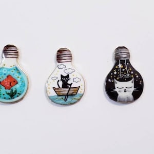 Pu&ograve; includere: Tre ornamenti in ceramica a forma di lampadina con disegni dipinti a mano. Il primo ornamento presenta un pesce rosso in un oceano blu e verde. Il secondo ornamento presenta un gatto nero in una barca con nuvole nel cielo. Il terzo ornamento presenta un gatto bianco con stelle in un cielo nero.