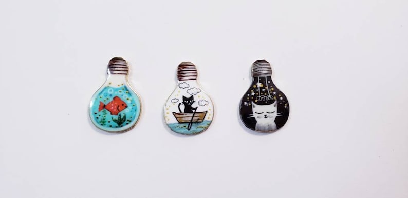 Pu&ograve; includere: Tre ornamenti in ceramica a forma di lampadina con disegni dipinti a mano. Il primo ornamento presenta un pesce rosso in un oceano blu e verde. Il secondo ornamento presenta un gatto nero in una barca con nuvole nel cielo. Il terzo ornamento presenta un gatto bianco con stelle in un cielo nero.