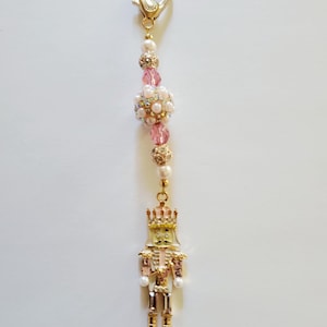 May include: A gold-toned charm with a lobster clasp, featuring a beaded strand with pink and pearl accents, and a miniature nutcracker figure. The nutcracker is detailed with gold, pink, and white enamel.
