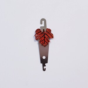 May include: A silver crochet hook with a decorative orange leaf charm. The hook has a loop at the top for hanging.