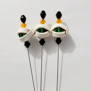 May include: Set of three white and green mummy eye pins with black and orange beads. The pins are attached to silver metal stems.