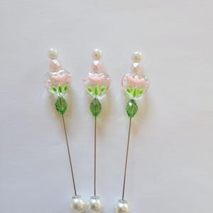 May include: Three decorative hat pins with floral designs. Each pin features a clear glass flower with pink petals, green leaves, and a pearl accent. The pins have a silver-colored stem and a large white pearl at the base.