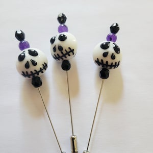 May include: Three decorative pins with white skull beads, black accents, and purple beads. The pins have silver-colored shafts and are arranged on a white surface. The skull beads have black eyes and stitched mouths.