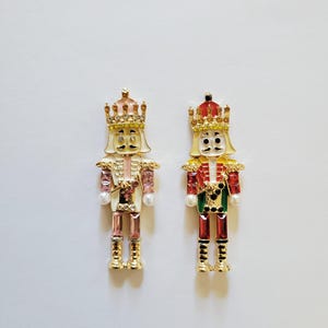 May include: Two decorative nutcracker figurines. One is in pink and gold, the other in red and gold. Both have gold crowns and are embellished with rhinestones and pearls. The figurines are on a white background.