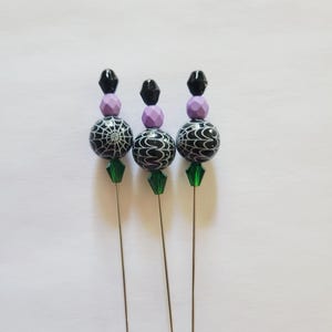 May include: Three decorative hat pins with black and white spiderweb and swirl patterned beads. Each pin has a lavender and black bead accent, and a green crystal. The pins have silver-colored metal shafts and end caps.