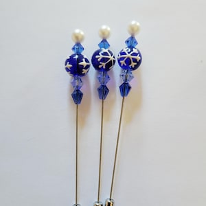 May include: Three decorative hat pins. Each pin features a dark blue glass bead with a white snowflake design, blue crystal accents, and a faux pearl top. The pins have silver-colored metal shafts and end caps.