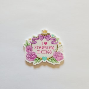May include: A die-cut sticker featuring an embroidery hoop design with the text I heart STABBING THINGS in pink cross-stitch font. The hoop is decorated with a purple ribbon, a gold crown, pink roses, green leaves, and a small teal heart.
