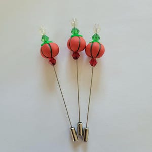 May include: Three orange and green pumpkin-shaped pins with silver stems. The pins are on a white background.