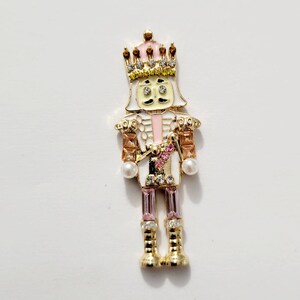 May include: A gold-colored nutcracker figurine with a pink crown, white face, and pink and brown accents. The figurine is decorated with rhinestones and pearls.