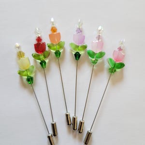 May include: Six colorful flower pins with green leaves and clear crystal accents. The pins are made of glass beads and have silver metal stems.