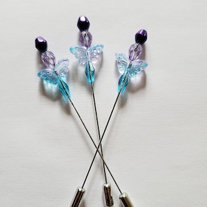 May include: Three decorative hat pins with butterfly designs. Each pin features a silver-colored base, a long, thin metal shaft, and a beaded top. The beads are purple, blue, and clear, creating a whimsical and elegant look.