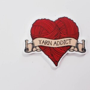 May include: A heart-shaped sticker with a red yarn ball design. A banner across the heart reads "YARN ADDICT" in black text. The sticker has a white border and is set against a white background.