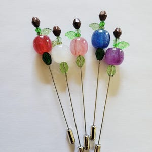 May include: A collection of six hat pins with colorful, round, bead-like tops. Each pin features a different colored bead, including red, white, pink, blue, and purple. The pins have silver-colored stems and decorative accents.