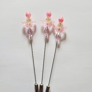 May include: Three decorative hat pins with pink and clear butterfly-shaped beads. Each pin has a silver-colored metal shaft and a cylindrical base. The beads are arranged in a layered design.
