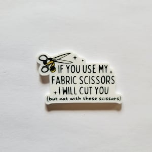 May include: A white die-cut sticker featuring a black illustration of scissors wrapped in a yellow measuring tape. The text reads: IF YOU USE MY FABRIC SCISSORS I WILL CUT YOU (but not with these scissors) in a black, hand-lettered style font.
