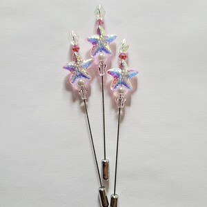 May include: Three star-shaped hat pins with iridescent, glittery surfaces. Each pin features a star, a pearl, and a clear crystal bead. The pins have silver-colored metal shafts and end caps. The pins are arranged on a white background.