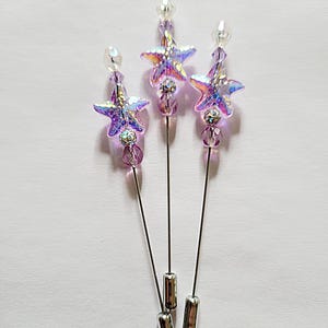 May include: Three decorative hat pins with iridescent star-shaped beads. Each pin features a star, a cluster of purple beads, and a clear bead at the top. The pins have silver-colored metal shafts and end caps. The background is a plain white surface.
