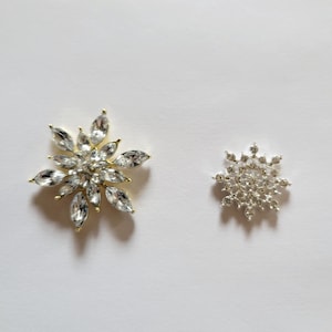 May include: Two decorative snowflake-shaped brooches. One is gold with clear rhinestones and the other is silver with clear rhinestones.