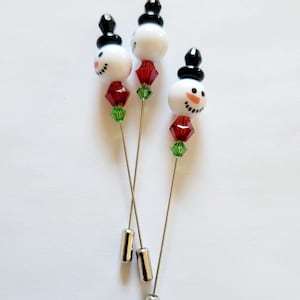 May include: Three snowman pins with black hats, white faces, red noses, and green and red beads. The pins are silver and have a silver cap at the bottom.