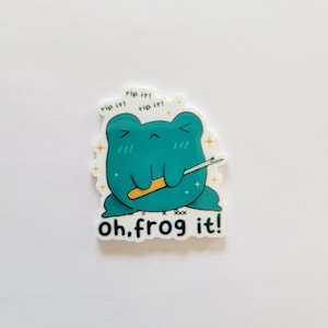 May include: A teal frog sticker with the text "rip it! rip it! rip it!" above it and "oh, frog it!" below it. The frog is holding a needle and thread.
