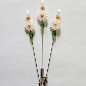 May include: Three silver-toned metal hat pins, each topped with a white daisy-shaped bead featuring an orange center. Below the flower is a green faceted bead, and above it is a small orange crystal bead topped with a clear, pointed bead.