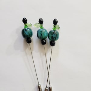 May include: Three decorative hat pins with a teal and black bead design. Each pin features a teal, round bead with a marbled effect, accented by black and green beads. The pins have silver-colored metal shafts and tips, set against a white background.