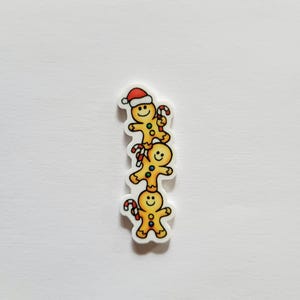 May include: A stack of three gingerbread men with smiling faces and candy canes. The top gingerbread man wears a red Santa hat. The gingerbread men are yellow with brown outlines and are on a white background.