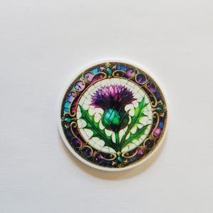 May include: Round coaster featuring a stained-glass design of a purple thistle flower with green leaves. The coaster has a white background and a decorative border with blue, purple, and gold accents. The coaster is a decorative item.