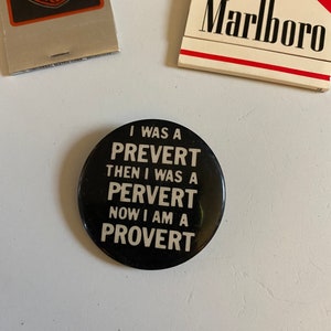 May include: A black button with white text that reads "I was a Prevert then I was a Pervert now I am a Provert". The button is on a white surface. There are two matchbook covers in the background, one with the Harley-Davidson logo and the other with the Marlboro logo.