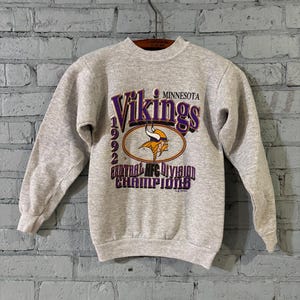 May include: A heather gray sweatshirt featuring the text "Vikings Minnesota 1992 Central AFC Division Champions" in purple and gold. The sweatshirt has a crew neck and long sleeves, and is hanging on a wooden hanger.