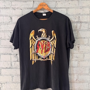 May include: Black t-shirt featuring a gold and red eagle graphic with the word "SLAYER" in red. The shirt is hanging on a wooden hanger against a white brick wall.