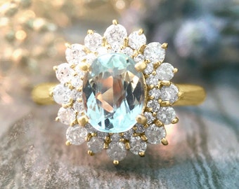 May include: A gold ring featuring a large, oval-cut, light blue gemstone at the center, surrounded by a halo of numerous small, round, clear white stones. The setting is crafted from yellow gold, with the stones held in place by small metal prongs.