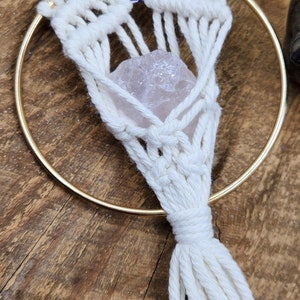 May include: A white macrame wall hanging with a pink crystal and a blue evil eye charm. The hanging is attached to a gold hoop.