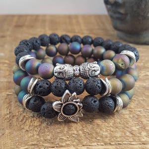 May include: Three layered bracelets featuring black lava rock beads, iridescent agate beads, and silver charms. The charms include two Buddha faces and a flower.