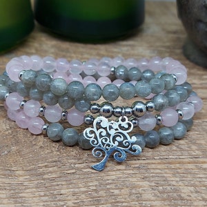 May include: A set of three bracelets featuring grey and pink beads with a silver tree charm. The bracelets are stacked on top of each other.