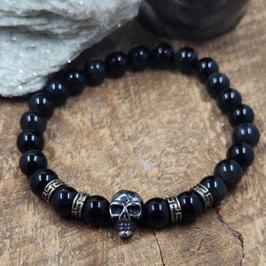 May include: Black beaded bracelet with a silver skull charm and gold accents.