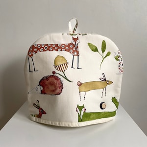 Puede incluir: Una funda para tetera blanca con un estampado de animales del bosque de estilo caprichoso. La funda presenta zorros, erizos, conejos y bellotas en tonos marrones, rojos, verdes y amarillos.