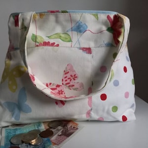Puede incluir: Una bolsa de tela blanca con estampado floral y de mariposas. La bolsa tiene un solo asa y un pequeño bolsillo en la parte delantera. La bolsa está sobre una superficie blanca con algunas monedas y billetes.
